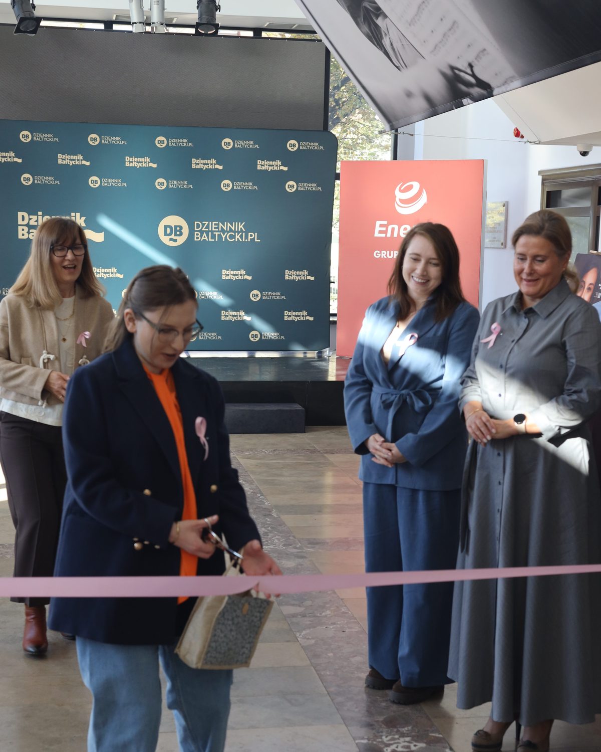 Klaudia Kozak przecina nożyczkami różową wstęgę na otwarciu wystawy „KobieTy to siła”, za nią stoją Małgorzata Kozłowska i Magdalena Raba